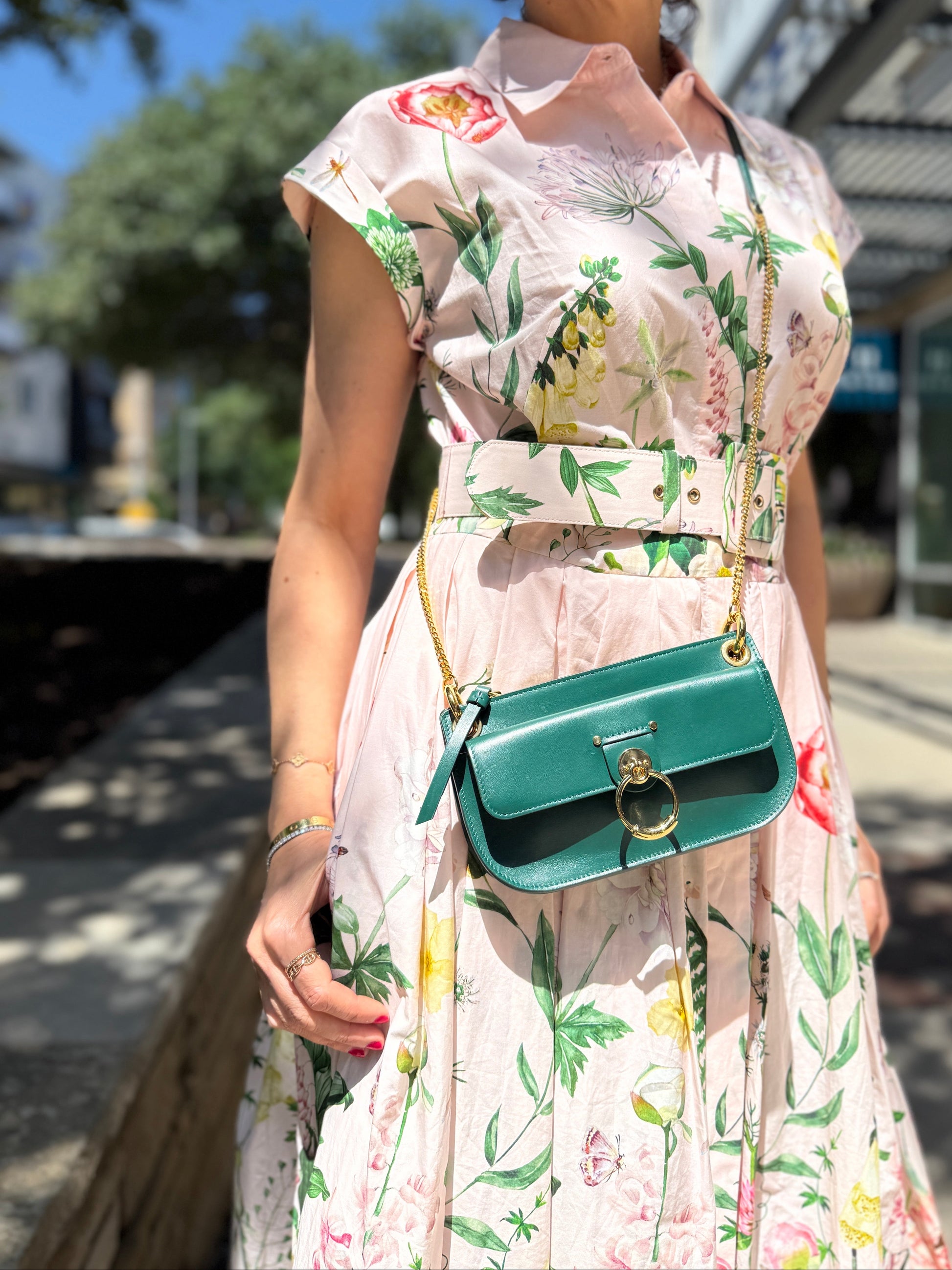 Woman in a floral dress holding a green handbag outdoors.